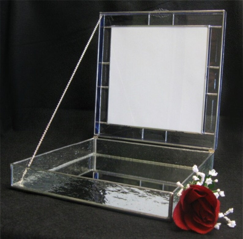 May include: A clear glass box with a hinged lid and a silver chain. The lid has a stained glass frame with a white background. The box is open and the inside is lined with a mirror. A red rose with white flowers is sitting on the box.