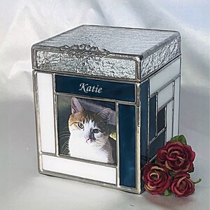 May include: A white and blue stained glass pet urn with a photo of a cat and the name "Katie" inscribed on the front.