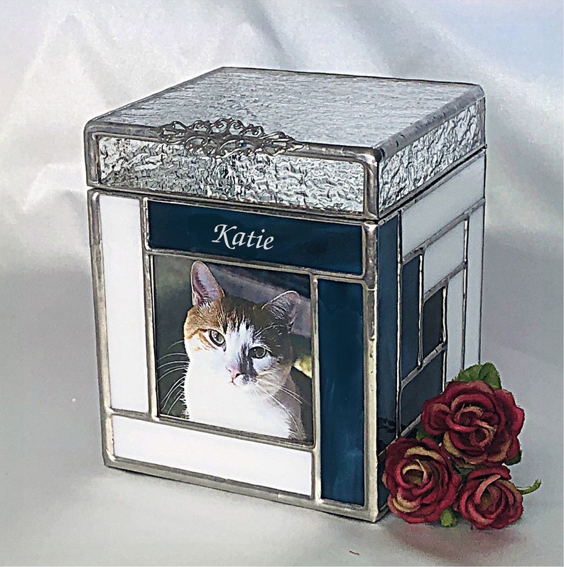 May include: A white and blue stained glass pet urn with a photo of a cat and the name "Katie" inscribed on the front.