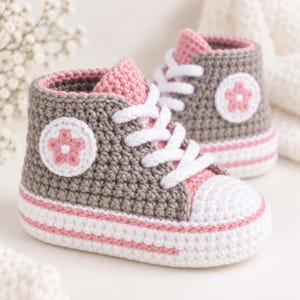 May include: A pair of high-top trainers crocheted with grey yarn, featuring white laces, a white toe cap, and a pink trim along the sole. Each shoe displays a circular white patch on the side with a pink five-petaled flower design in the centre.
