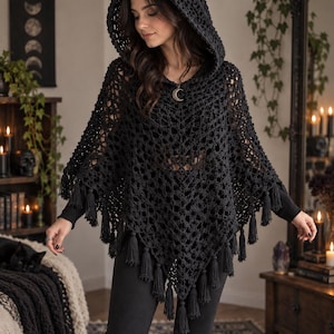 May include: A black crochet poncho featuring an openwork diamond pattern, a hood with pointed cat-like ears, and a hemline finished with multiple hanging tassels. The garment is worn over a black long-sleeved top and black trousers.