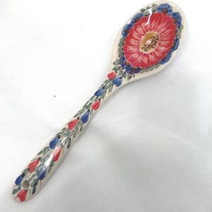 May include: A ceramic spoon with a cream-colored base featuring a large red flower in the bowl and a pattern of small red and blue floral buds with green leaves extending down the handle, which includes a small circular hole at the end.