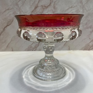 May include: A clear glass pedestal bowl featuring a vibrant red rim. The bowl has a wide, circular opening, a textured pattern of raised dots around the midsection, and a sturdy, decorative base with a matching embossed design.