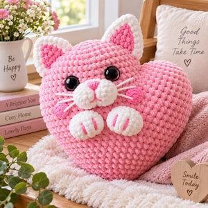 May include: A heart-shaped plush cat toy made of chunky pink yarn with white accents on the ears, muzzle, and paws. The cat features large black plastic eyes, a pink stitched nose, and white whiskers. The texture is soft and crocheted.