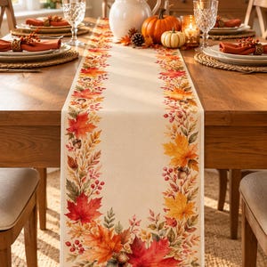 May include: A rectangular cream-colored table runner featuring a printed border of vibrant autumn foliage, including red and orange maple leaves, small brown acorns, and clusters of red berries, extending along the sides and bottom edges.