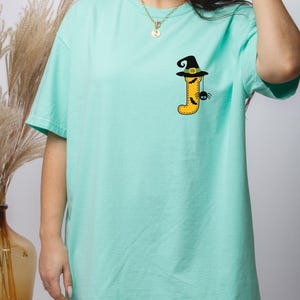 May include: A mint green short-sleeve t-shirt featuring a graphic on the left chest. The graphic displays the letter J in yellow with a black witch hat on top, decorated with a green band, small black bats, and a dangling black spider.
