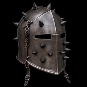 May include: A dark metallic medieval-style helmet featuring a pointed visor, multiple sharp conical spikes arranged across the surface, and a decorative metal chain attached to the side. The helmet includes leather chin straps with metal buckles.