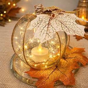 May include: A clear glass pumpkin-shaped container wrapped in thin twine, containing a battery-operated LED tea light and fairy lights. The top is decorated with a textured fabric maple leaf and a twine-wrapped stem, resting on a glass plate.