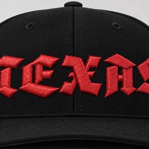 Könnte beinhalten: Eine schwarze Baseballkappe mit dem Wort TEXAS, das in fetten, roten Buchstaben im gotischen Stil auf die Vorderseite gestickt ist. Die Kappe hat eine strukturierte Krone, einen flachen Schirm und einen Knopf oben, was ein klares Design ergibt.