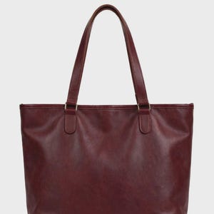 Peut inclure: Un sac cabas bordeaux profond à la finition lisse, doté de deux longues anses fixées par des attaches métalliques rectangulaires. Le sac présente une ouverture large sur le dessus avec des coutures renforcées aux coins de la base.