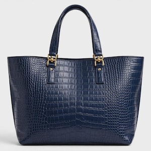 Peut inclure: Un sac cabas bleu foncé doté d'une texture imitation peau de crocodile. Le sac possède deux anses robustes fixées par des éléments métalliques dorés. Le design présente une large ouverture et une base rectangulaire structurée.