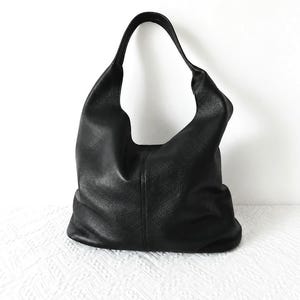 May include: A black hobo bag with a textured surface and a single integrated shoulder strap. The bag features a slouchy, unstructured silhouette with a vertical seam running down the center of the front panel, creating a simple and functional design.