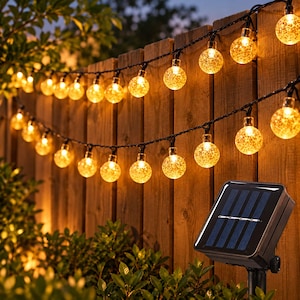Op de afbeelding: Een solar-lichtsnoer voor buiten met heldere, getextureerde bolvormige lampen die een warm geel licht uitstralen. De lampen hangen over een houten schutting, vergezeld door een zwart rechthoekig zonnepaneel op een paal.