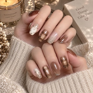 Può includere: Unghie curate con un mix di toni marroni e beige. I design includono motivi a quadri marroni, accenti di glitter dorati e piccoli punti metallici su una base chiara. Le unghie hanno una finitura lucida e bordi arrotondati.