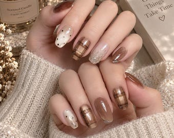 Pode incluir: Unhas manicuradas com uma mistura de tons castanhos e bege. Os designs incluem padrões de xadrez castanho, detalhes de glitter dourado e pequenos pontos metálicos sobre uma base clara. As unhas têm um acabamento brilhante e arredondado.