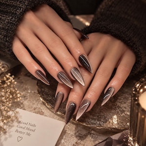 May include: Long, pointed stiletto-shaped nails featuring a dark brown and metallic silver cat-eye magnetic polish effect. A small white card is visible with the printed text: Good Nails, Good Mood, Better Me, followed by a small heart symbol.