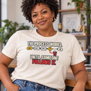 May include: A cream-colored short-sleeve t-shirt featuring a graphic of a flowchart with the text "THE PROCESS WORKS. UNTIL SOMEONE FOLLOWS IT." The flowchart includes boxes labeled "START," "STEP 1," "STEP 2," a question mark, and a trash can icon.