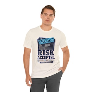 May include: A cream-coloured short-sleeve t-shirt featuring a graphic print of a document with the text ACCEPTED, RISK ACCEPTED, and a table detailing risk management metrics including system outage, IT operations, medium impact, and low likelihood.