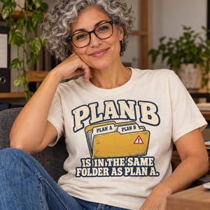 Op de afbeelding: Een crèmekleurig t-shirt met korte mouwen en een afbeelding van een gele map met tabbladen met het label Plan A en Plan B. De tekst luidt PLAN B IS IN THE SAME FOLDER AS PLAN A in donkerblauwe letters met een rood waarschuwingsdriehoekje.