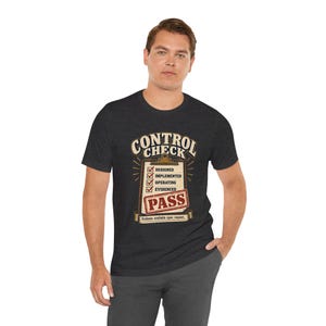 May include: A dark heather grey short-sleeve t-shirt featuring a graphic of a clipboard with the text CONTROL CHECK at the top. The list includes DESIGNED, IMPLEMENTED, OPERATING, and EVIDENCED with red checkmarks, and a red PASS stamp at the bottom.
