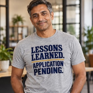 May include: A heather gray short-sleeve t-shirt featuring the dark blue text "LESSONS LEARNED." above a thin horizontal gold line, with the dark blue text "APPLICATION PENDING." printed directly below the line on the chest.
