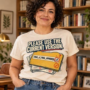 May include: A cream-colored short-sleeve t-shirt featuring a graphic of a yellow file folder stacked on papers. The text reads: PLEASE USE THE CURRENT VERSION. The folder is labeled FINAL_v2_FINAL_APPROVED with a red v7 circle and checkmark.