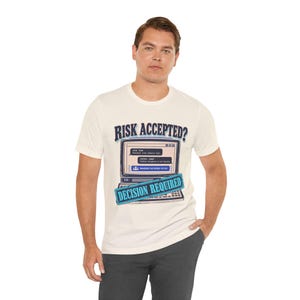 Puede incluir: Una camiseta de manga corta de color crema con un gráfico de un monitor de ordenador que muestra un cuadro de diálogo con el texto RISK ACCEPTED? y DECISION REQUIRED. La pantalla muestra mensajes técnicos sobre riesgos.