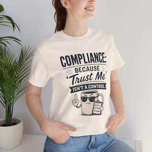 Può includere: Una t-shirt a maniche corte color crema con il testo blu scuro COMPLIANCE: BECAUSE 'TRUST ME' ISN'T A CONTROL. Sotto il testo c'è la grafica di una tazza da caffè beige con occhiali da sole neri che tiene in mano una cartellina.