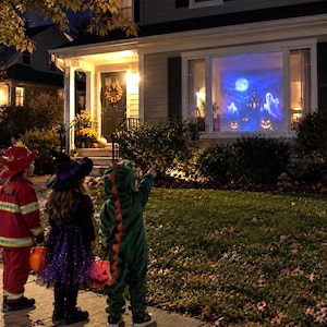 May include: A house window displays a blue-toned Halloween projection featuring a full moon, a haunted mansion, ghosts, and glowing jack-o'-lanterns. Three children in costumes stand on a pavement, looking toward the illuminated window display.