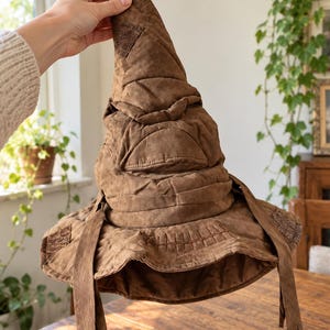 May include: A brown fabric hat with a tall, pointed, and folded crown. The hat features stitched details resembling a mouth and eyes, with a wide, floppy brim and two long, thin fabric strips hanging from the sides of the brim.