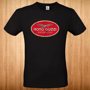 Op de afbeelding: Een zwart T-shirt met korte mouwen en een centraal ovaal motief met een verweerde rode achtergrond en een gouden rand. Het motief toont een gouden adelaarsymbool boven de witte tekst MOTO GUZZI MOTORCYCLES en de kleine tekst SINCE 1921.