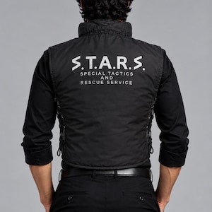 Puede incluir: Un chaleco táctico negro sobre una camisa negra de manga larga y pantalones negros. La parte posterior del chaleco presenta el texto blanco S.T.A.R.S. SPECIAL TACTICS AND RESCUE SERVICE. El chaleco tiene detalles de cordones laterales.