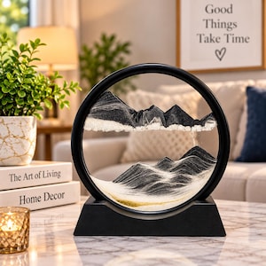 May include: A circular black frame containing a dynamic sand art display with layers of black, white, and gold sand. The sand forms mountain-like peaks and valleys within the glass enclosure, which is mounted on a solid black rectangular base.