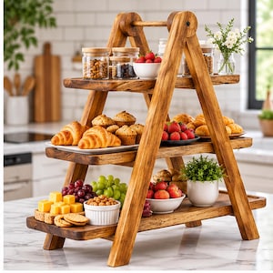 May include: A three-tiered wooden ladder-style serving stand featuring a natural wood grain finish. The stand holds various food items including croissants, muffins, strawberries, grapes, cheese cubes, crackers, and small glass jars.