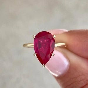 Op de afbeelding: Een ring met een peervormige, dieprode edelsteen gezet in een dunne, gepolijste gouden band. De steen is bevestigd met een zetting met meerdere pootjes en toont een levendige karmozijnrode kleur en een gefacetteerd oppervlak dat licht reflecteert.