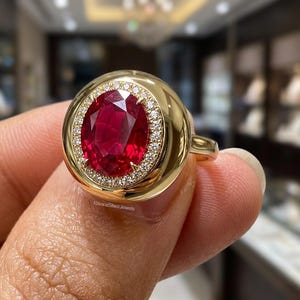 May include: A gold ring featuring a large, oval-cut, deep red gemstone at its center. The central stone is surrounded by a halo of small, clear, sparkling stones set within a smooth, polished gold frame. The text RivaraSilverJewels is visible.