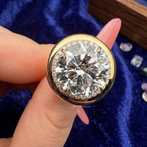 May include: A gold ring featuring a large, round, brilliant-cut clear stone at the center, surrounded by a halo of smaller, pavé-set clear stones. The metal band is smooth and polished, with the text RivaraSilverJewels visible on the setting.