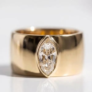Könnte beinhalten: Ein breiter Goldring mit einem einzelnen, im Marquise-Schliff facettierten, klaren Edelstein in der Mitte. Die polierte Metalloberfläche des Rings reflektiert das Licht und betont die glatten, abgerundeten Kanten der Ringschiene und den Stein.
