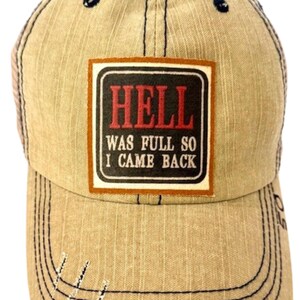 May include: A tan baseball cap with a distressed finish and dark stitching. A rectangular patch on the front features a black background with a white border and the red and white text: HELL WAS FULL SO I CAME BACK. The cap has a structured crown.