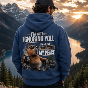 May include: A blue hooded sweatshirt featuring a graphic on the back of a capybara wearing sunglasses, sitting in water, and holding a mug labeled "DO NOT DISTURB." The text reads, "I'M NOT IGNORING YOU. I'M JUST PRIORITIZING MY PEACE."