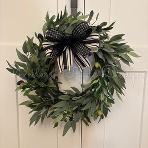 May include: A circular wreath featuring green olive branches with small dark and light green olives and white floral accents. A large decorative bow with black and white striped and checkered patterns is attached to the top of the foliage.