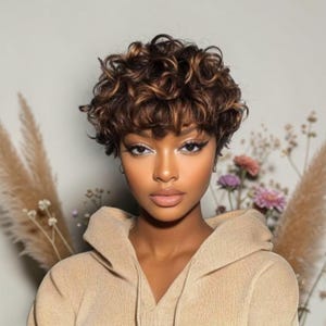 May include: A short, curly brown wig with warm highlights, featuring a textured, layered cut with soft curls framing the face. The hair has a mix of dark brown and lighter caramel tones, styled in a voluminous, rounded shape with a fringe.