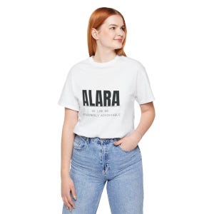 May include: A white short-sleeved t-shirt featuring the black text ALARA printed in a bold sans-serif font, with the smaller text AS LOW AS REASONABLY ACHIEVABLE printed directly underneath it, paired with light blue denim jeans.