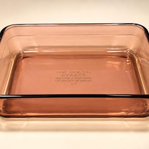 Puede incluir: Una fuente de horno rectangular de vidrio rosa transparente con esquinas redondeadas. La base tiene grabado el texto: 3.0 QT, 936 ML, 2.8 L, PYREX, FOR OVEN & MICROWAVE, NO STOVETOP OR BROILER, A-2. El recipiente presenta un acabado liso y uniforme.