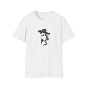 May include: A white short-sleeved t-shirt featuring a centered graphic illustration of a figure with long flowing hair wearing a wide-brimmed black hat with a thin gold band. The design is rendered in black line art with gold accents.