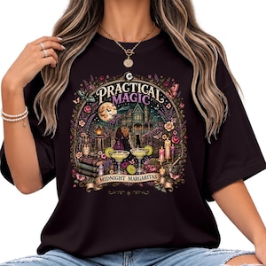 May include: A black t-shirt featuring a colorful graphic print with the text "PRACTICAL MAGIC" and "MIDNIGHT MARGARITAS". The design depicts a large house, two figures, a full moon, margaritas, books, candles, and flowers in a detailed illustration.