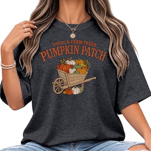 May include: A dark gray short-sleeve t-shirt featuring a graphic of a wooden wheelbarrow filled with pumpkins and autumn leaves. The text above the graphic reads "LOCAL & FARM FRESH PUMPKIN PATCH" in orange lettering. The design includes orange, green, and white pumpkins.