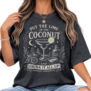 May include: A charcoal gray short-sleeve t-shirt featuring a white graphic print of a cocktail glass, a bottle labeled Witches Spirits, and the text Put the lime in the coconut and drink it all up Midnight Margaritas with celestial accents.
