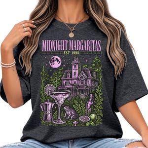 May include: A dark gray t-shirt featuring a graphic print with the text "MIDNIGHT MARGARITAS EST. 1998". The design includes a large house, a full moon, a margarita glass, a frog, a coffee pot, a bottle, and lime slices in shades of pink and green.