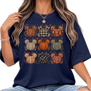 May include: A navy blue short-sleeve t-shirt featuring a grid of nine pumpkin illustrations, each with mouse-ear shapes. The pumpkins display various patterns including leopard print, black and white plaid, floral, polka dots, and solid orange.